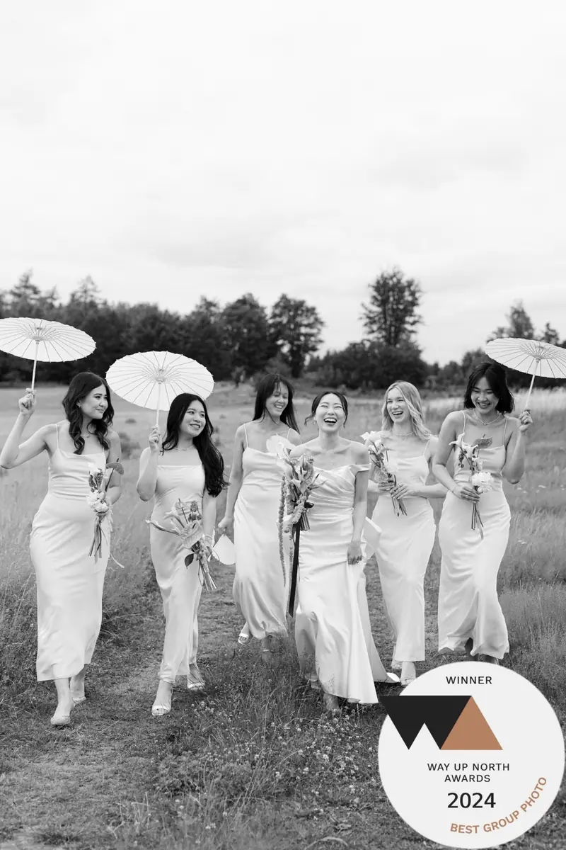 Black and white photo of a bride and her bridesmaids laughing in a meadow under white paper parasols, with a Way Up North Awards 2024 winner badge