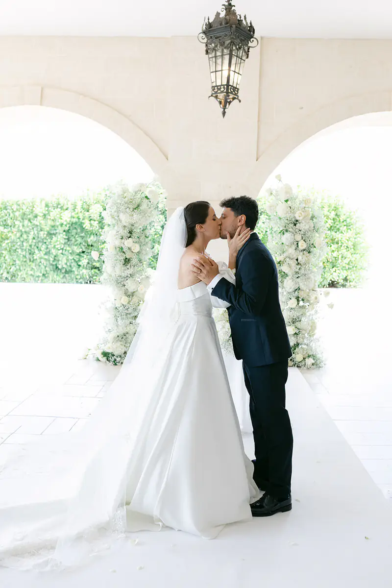 Bride and groom kissing under a white arcade decorated with flowers in Bari, Italy