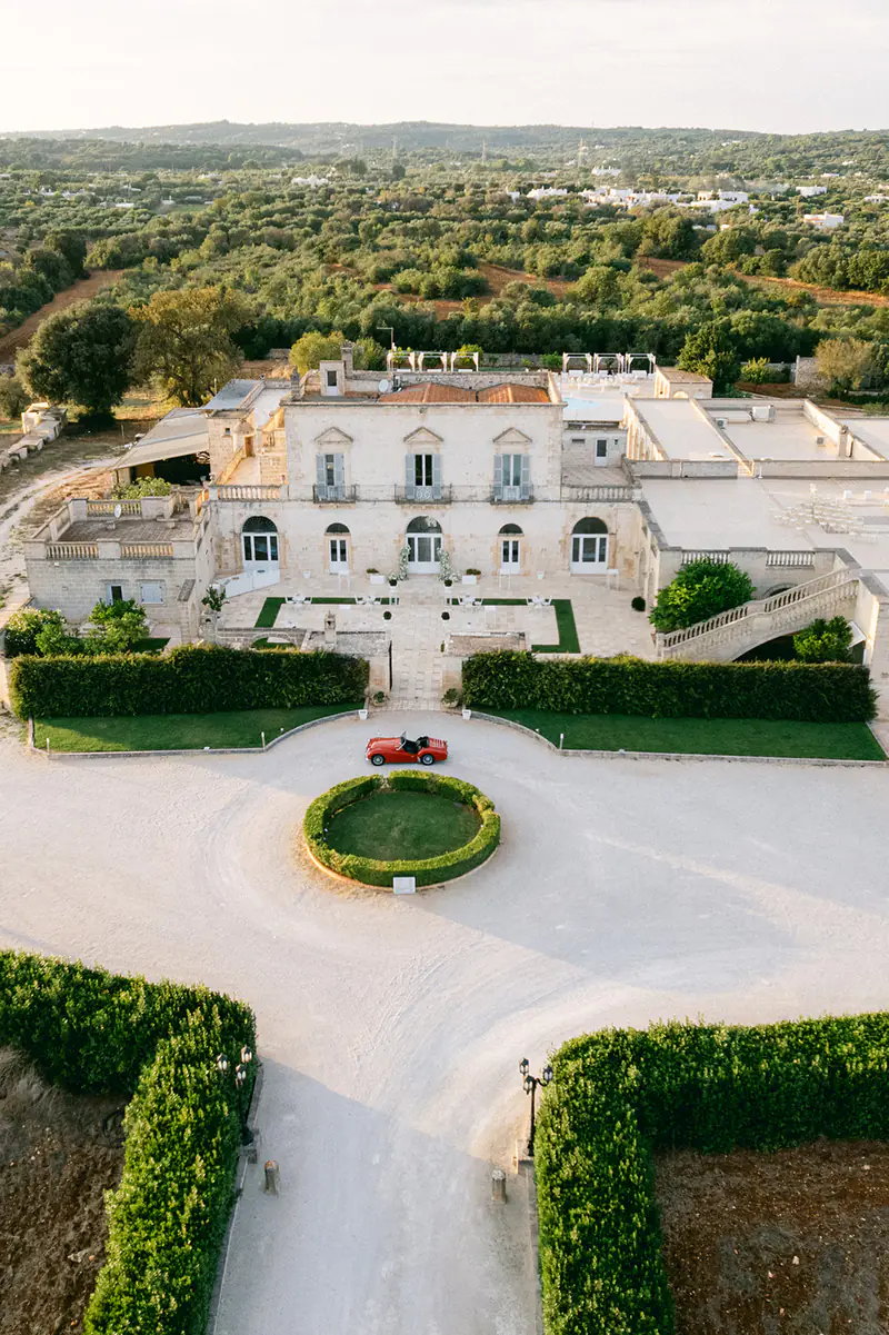 Aerial view of a white wedding villa with a red vintage car on the round drive, surrounded by olive groves