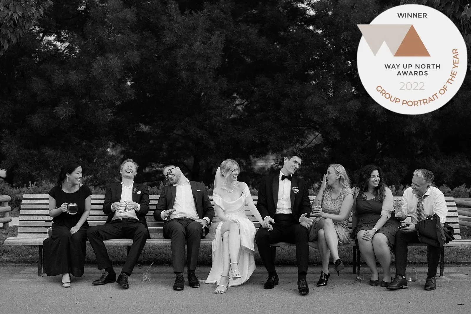Black and white group photo of the bride, groom and friends laughing on a park bench, with a Way Up North Awards 2022 badge for group portrait of the year