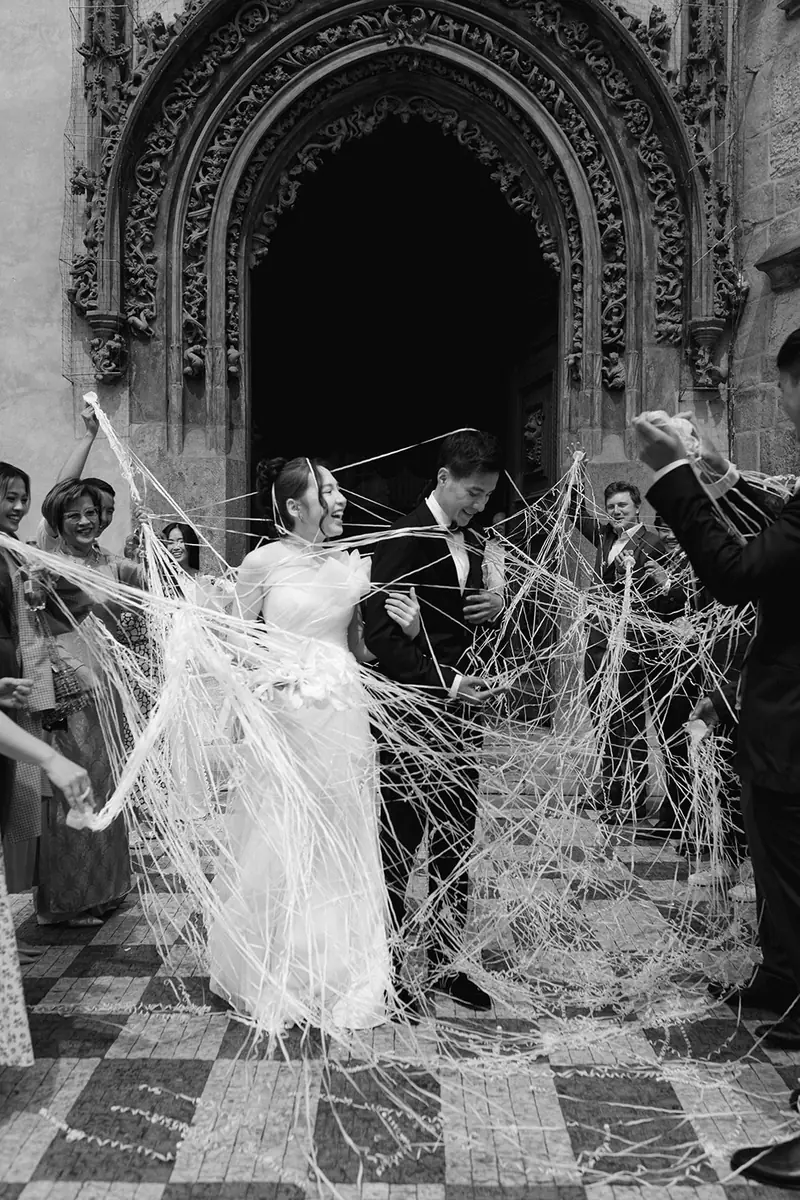 Black and white photo of a bride and groom walking out of a gothic doorway through a shower of streamers