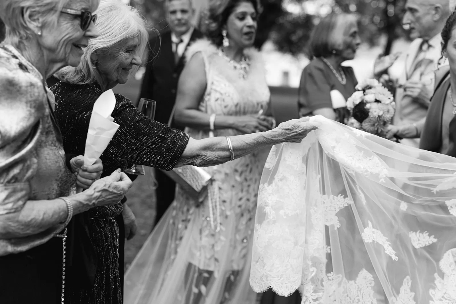 Black and white photo of smiling older guests reaching out to touch the bride’s long lace veil