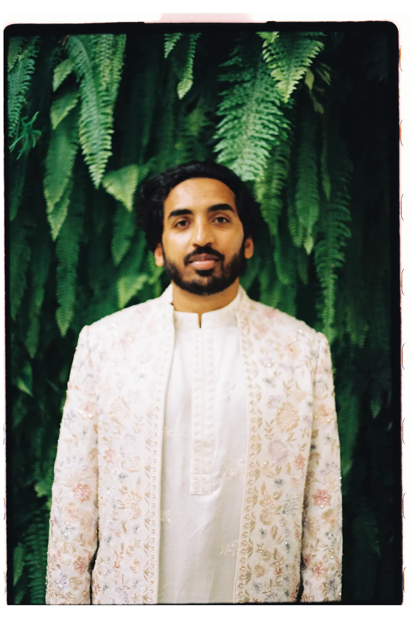 Groom in an embroidered ivory sherwani in front of a wall of ferns