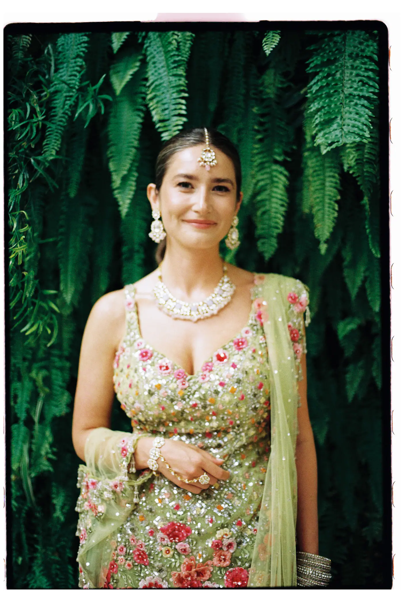 Bride in a pastel green embroidered lehenga and jewellery in front of a wall of ferns