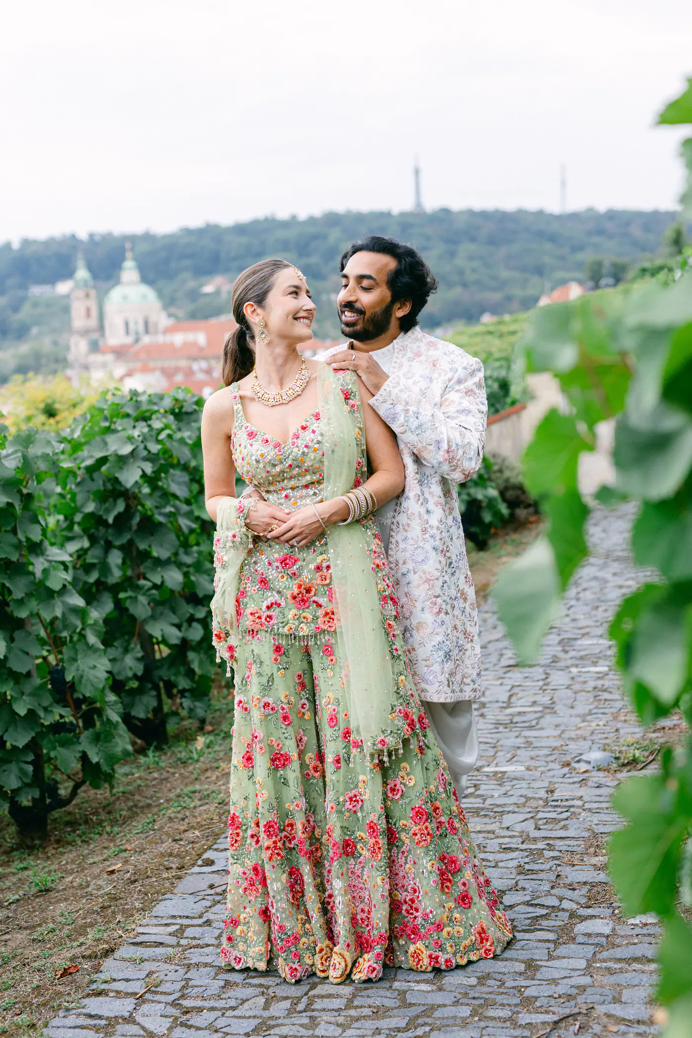 Bride and groom embracing on a cobbled path through the vineyard, with Prague behind them