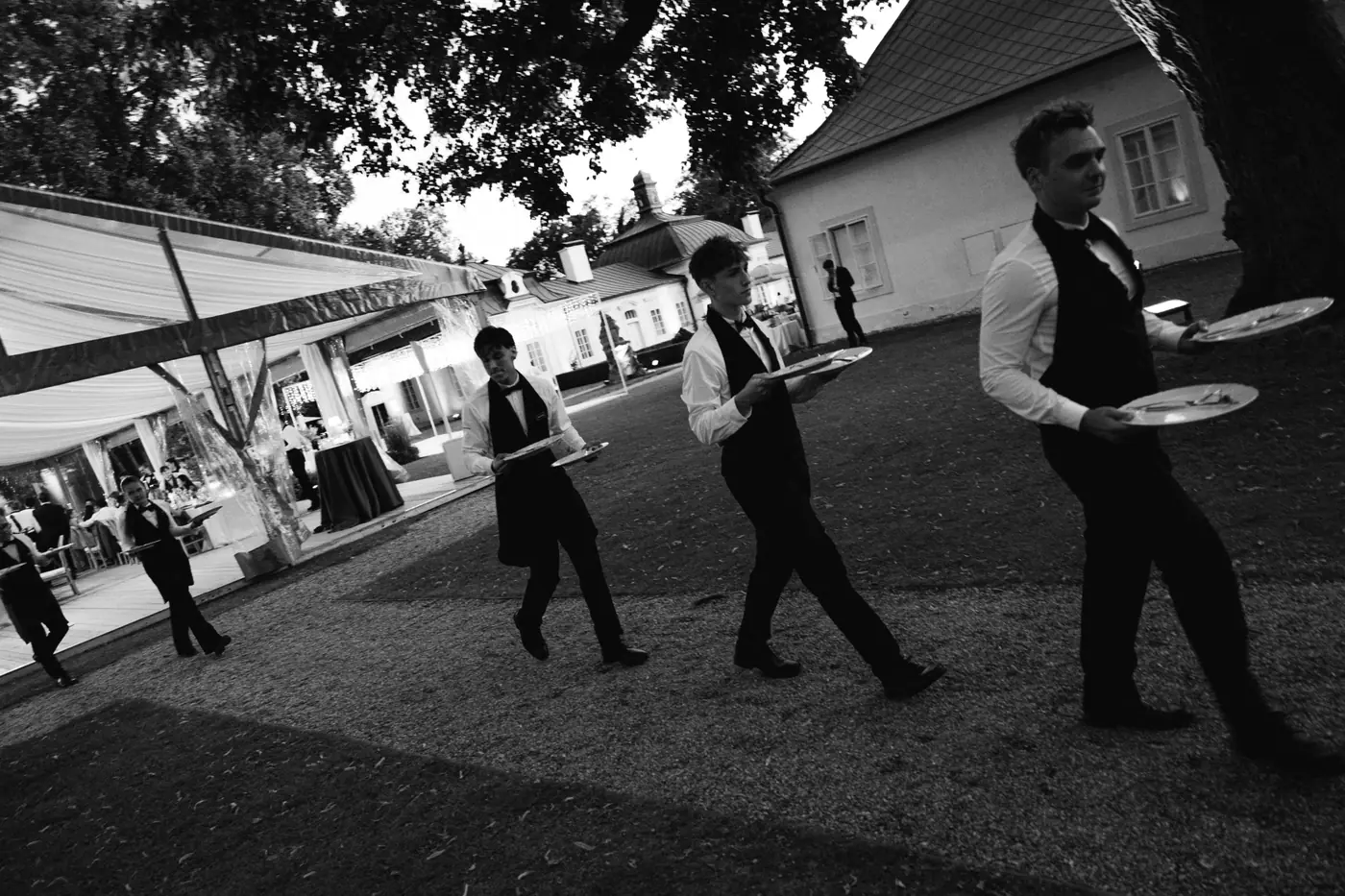 Black and white photo of waiters carrying trays across a gravel courtyard