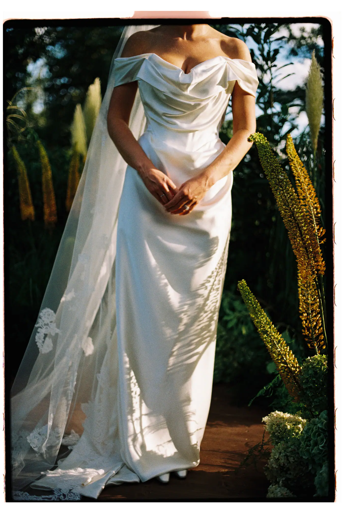 Bride in an off-shoulder satin dress with a long veil among tall yellow flowers