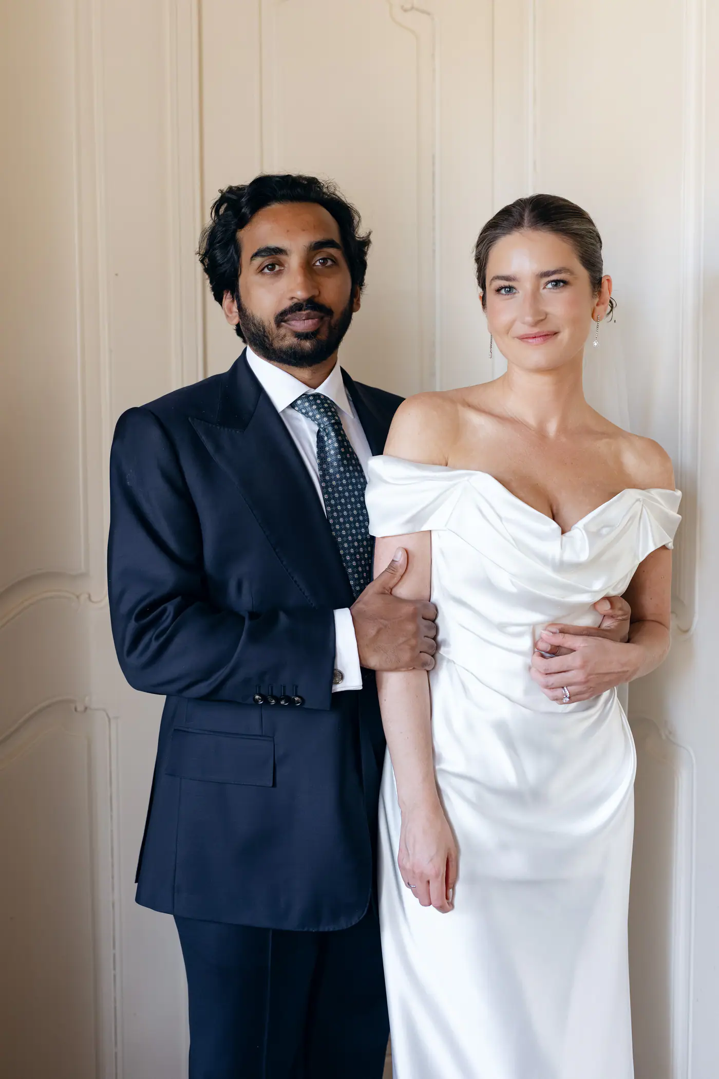 Bride and groom standing arm in arm against a white wall