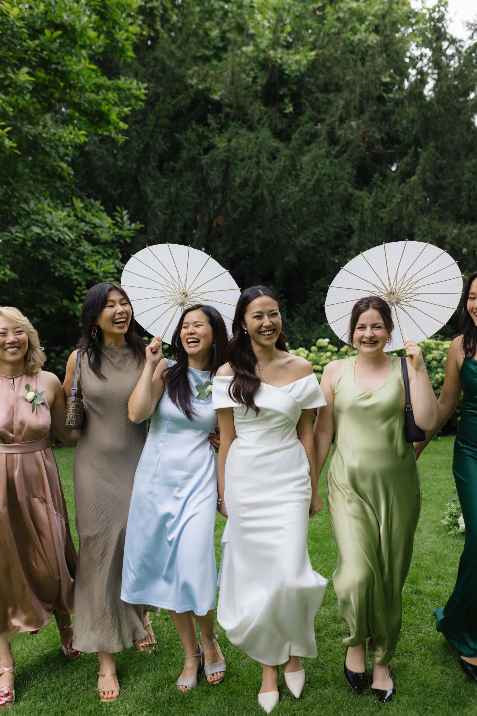 Bride and bridesmaids in pastel satin dresses holding white paper parasols in a garden