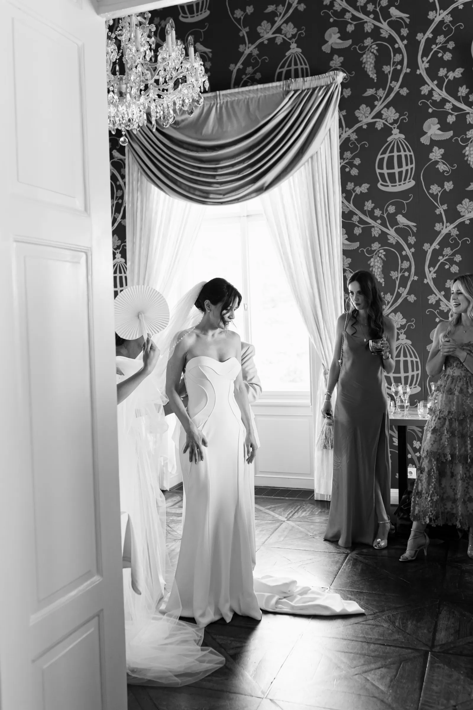 Black and white photo of a bride getting ready in a room with ornate wallpaper and a chandelier