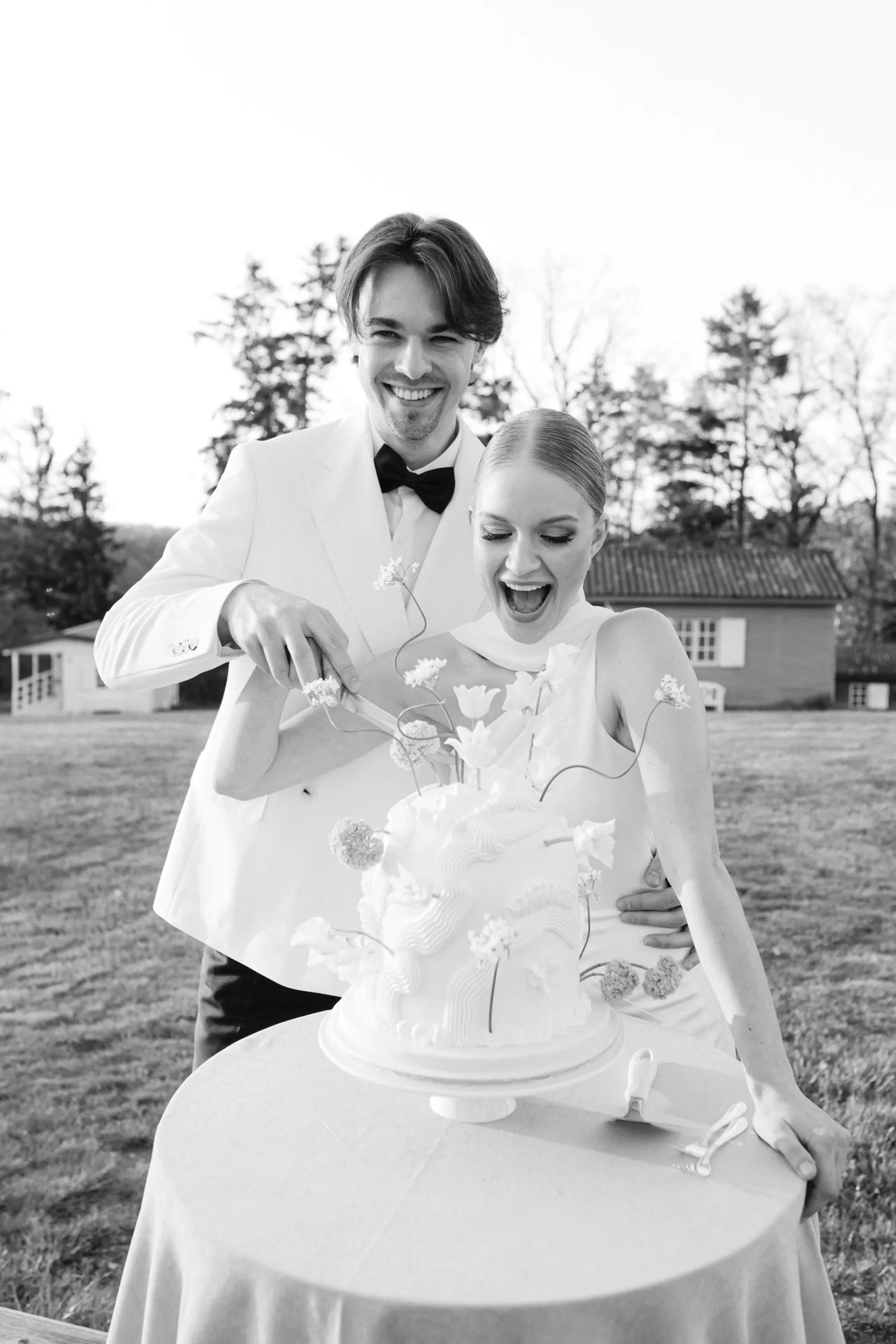Black and white photo of bride and groom cutting a sculptural wedding cake outdoors