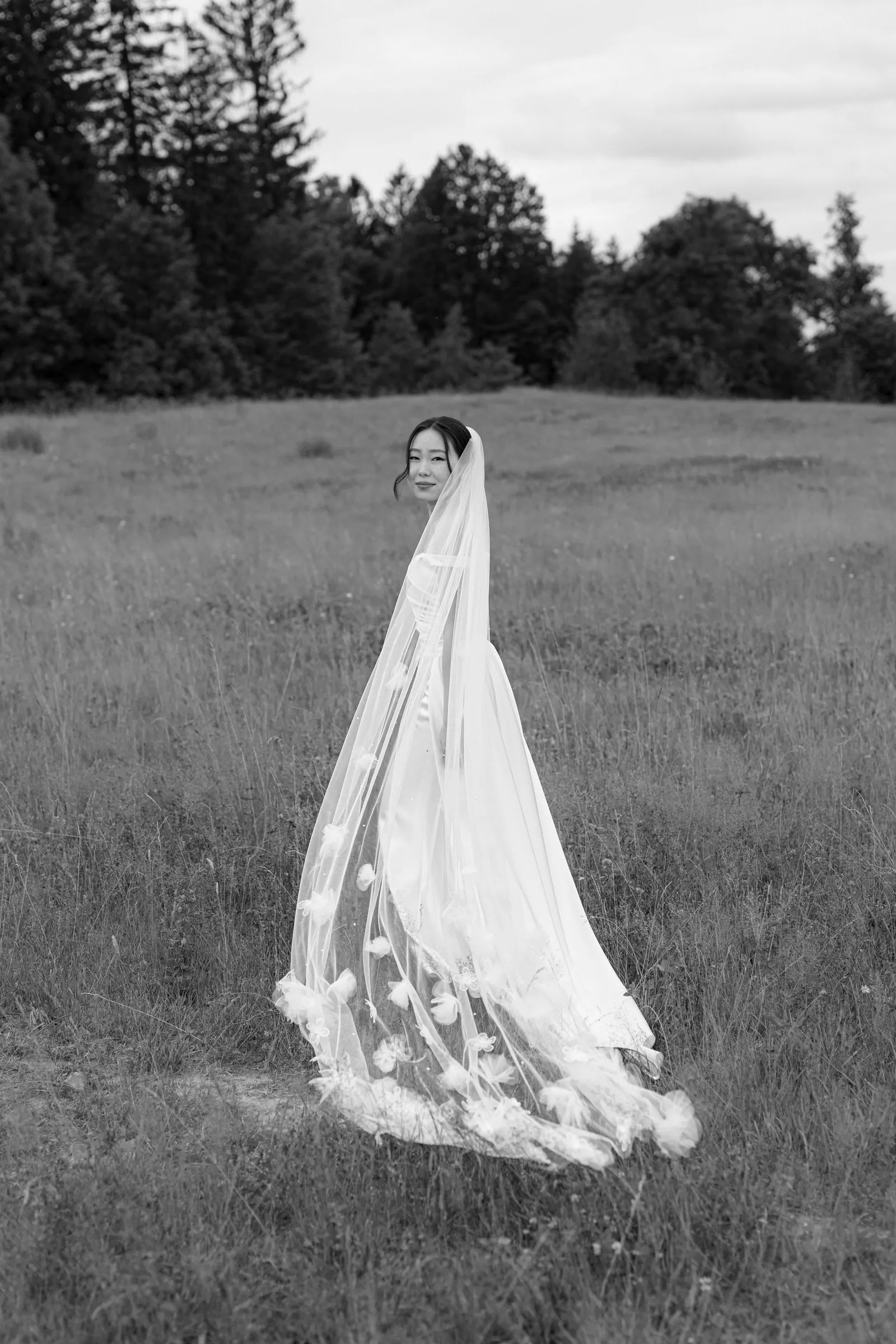 Black and white photo of a bride with a long veil walking through a meadow