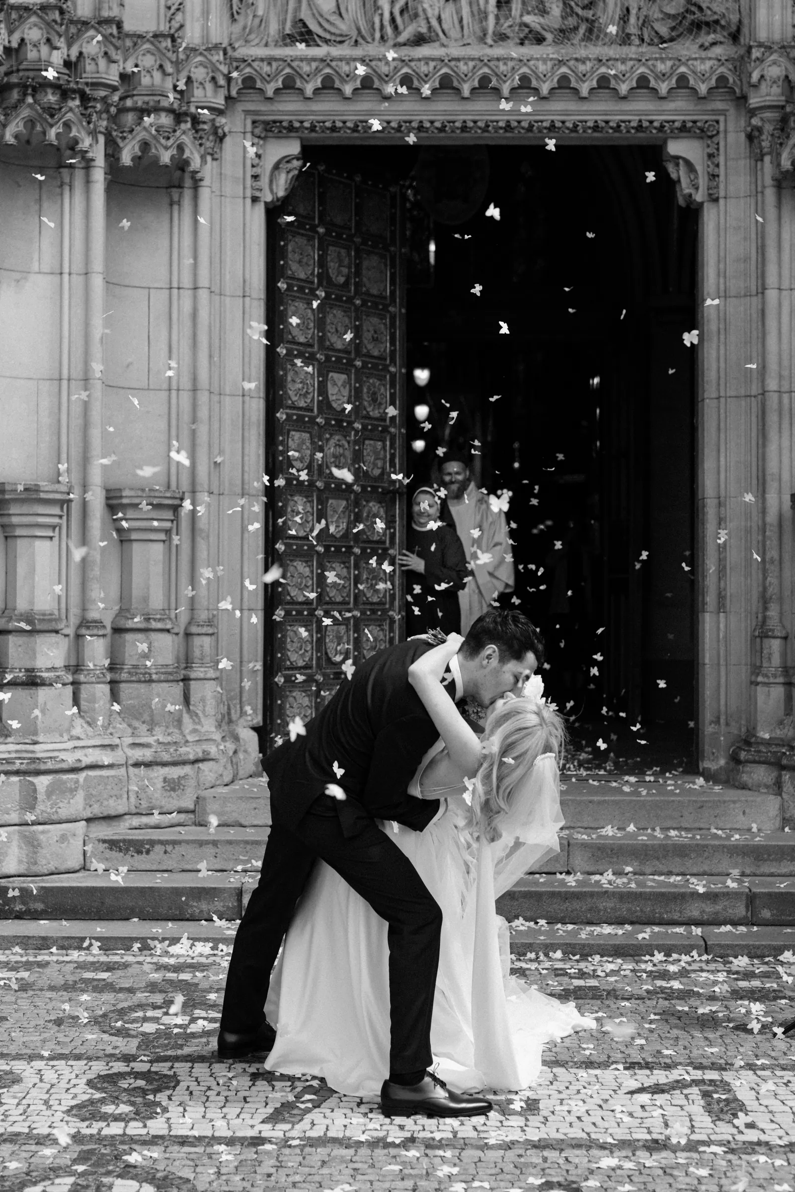Black and white photo of a couple kissing under confetti in front of a gothic church portal