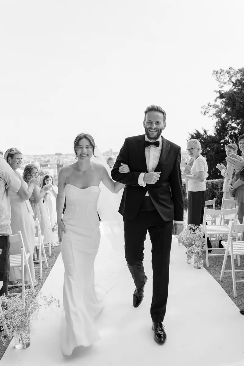 Black and white photo of bride and groom walking down a white aisle as guests applaud — Villa Richter, Prague