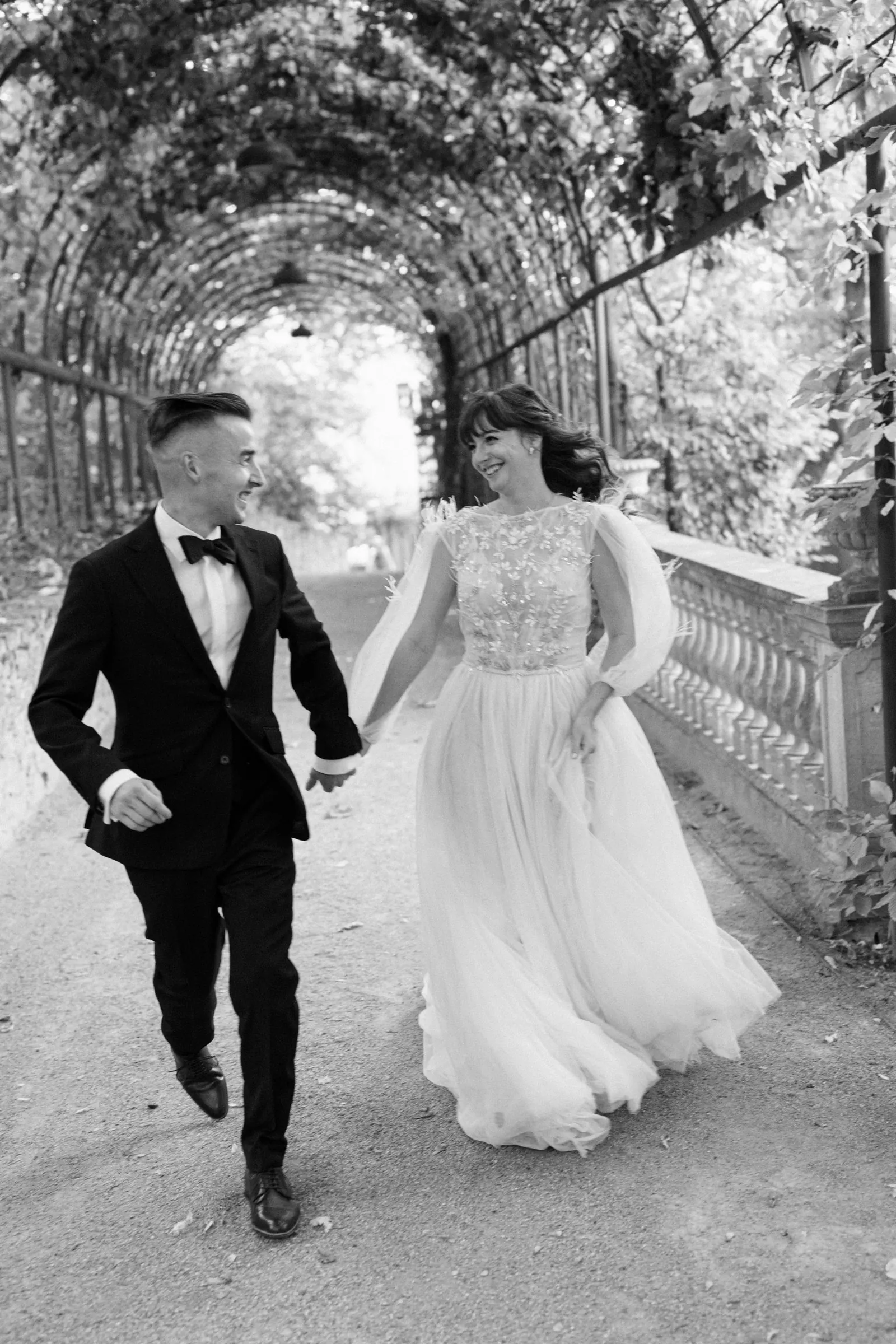 Black and white photo of bride and groom running hand in hand along a balustrade — Savoia Castle