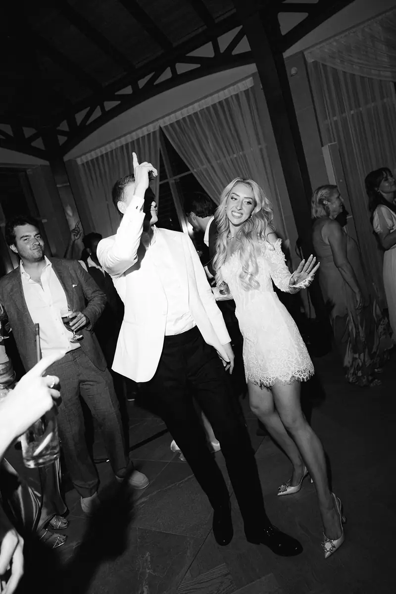 Black and white photo of the groom in a white jacket and the bride in a short lace dress dancing among their guests
