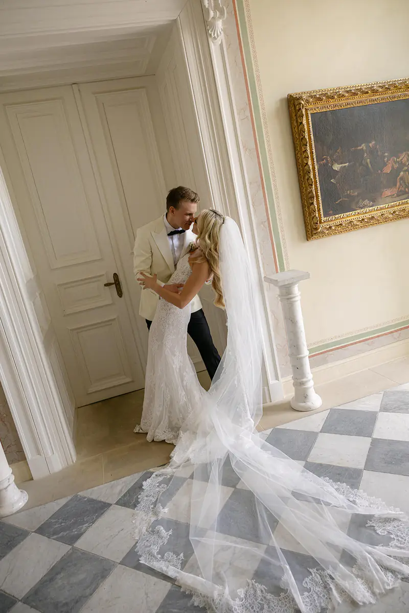Bride and groom kissing in a palace hallway with a checkerboard floor and a long lace veil